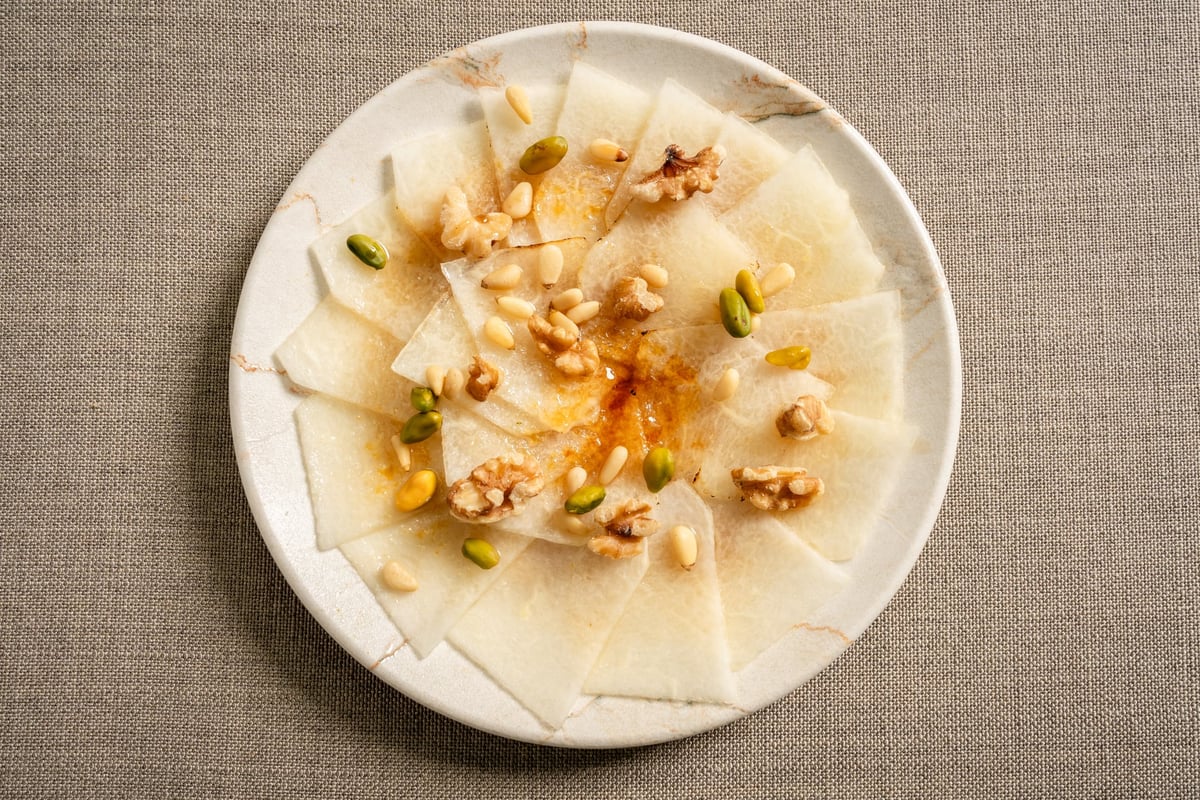Assiette de carpaccio de melon présentée sur un fond en tissu gris, accompagnée de morceaux d'ananas frais, de noix