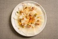 Assiette de carpaccio de melon présentée sur un fond en tissu gris, accompagnée de morceaux d'ananas frais, de noix