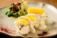 Plato de merluza cocinada con una delicada salsa pilpil de cítricos, decorado con finas láminas de limón amarillo.
